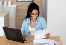 A woman with dark brown hair wearing a blue button-up shirt looking at paperwork and a laptop computer