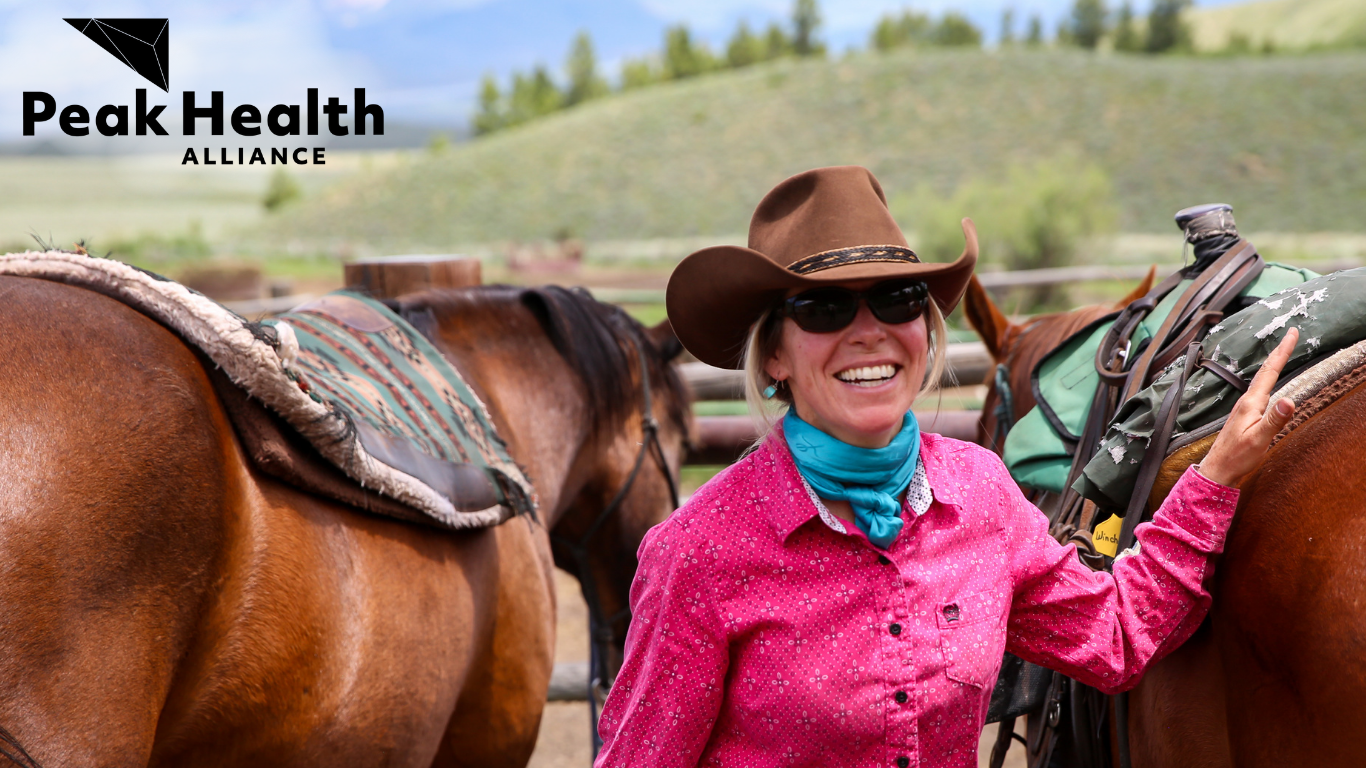 A woman in a pink hat with a teal scarf and cowboy had standing between two large brown horses. The Peak Health Alliance logo is in the top left corner.